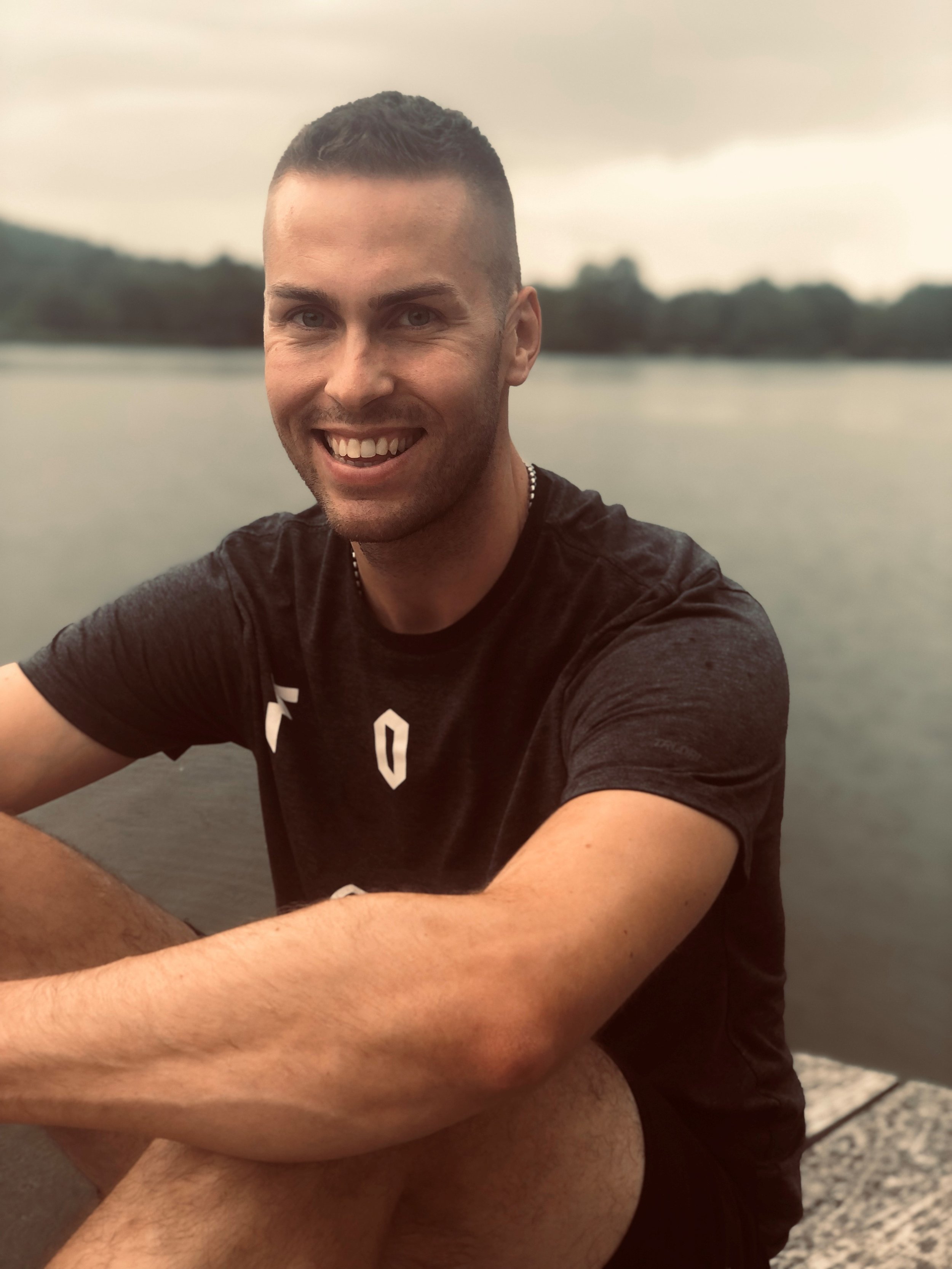 Lächelnder junger Mann mit dunklen Haaren, kurz geschnitten, fotografiert an einem See bei bewölktem Himmel, trägt ein schwarzes T-Shirt und sitzt auf einem Steg.