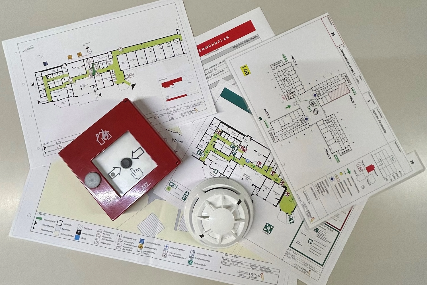 Verschiedene Gebäudepläne, Plakate und Materialien für Brandschutz auf einem Tisch, inklusive rotem Feueralarmschalter und Rauchmelder.