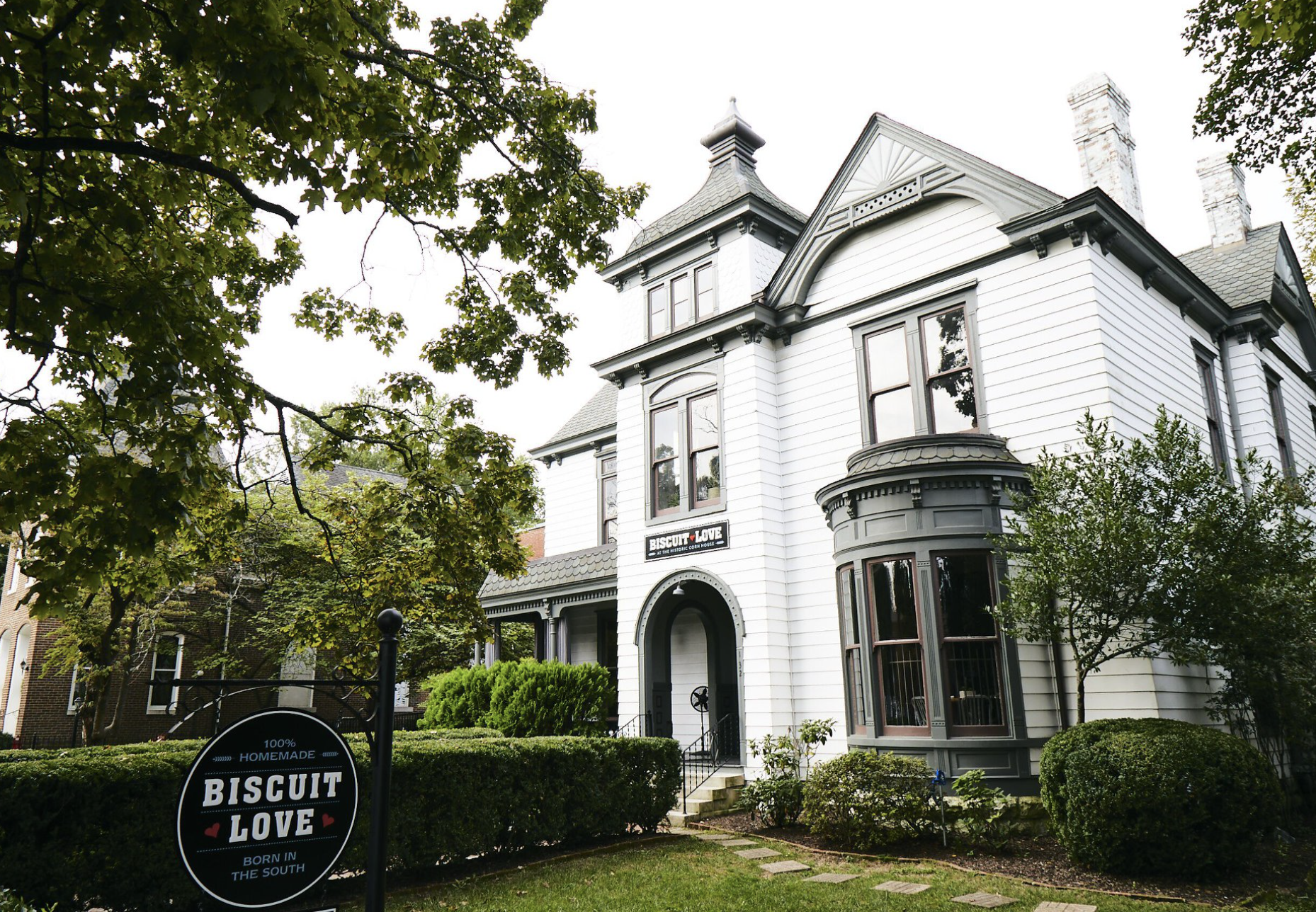 A white Victorian-style house with a rounded bay window and decorative trim, surrounded by green trees and shrubs, with a sign in the yard that reads 'Biscuit Love - Homemade, Born in the South'.