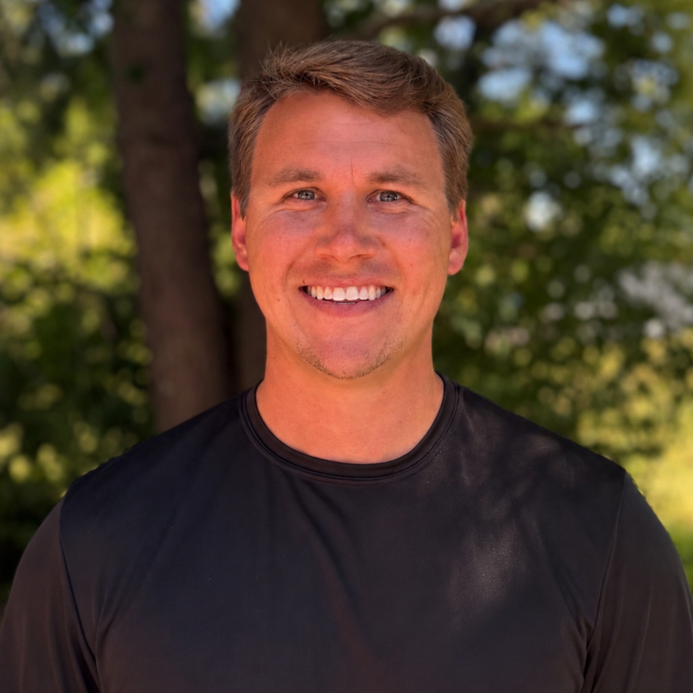 A smiling man with short light brown hair, wearing a black athletic shirt, outdoors with trees in the background.