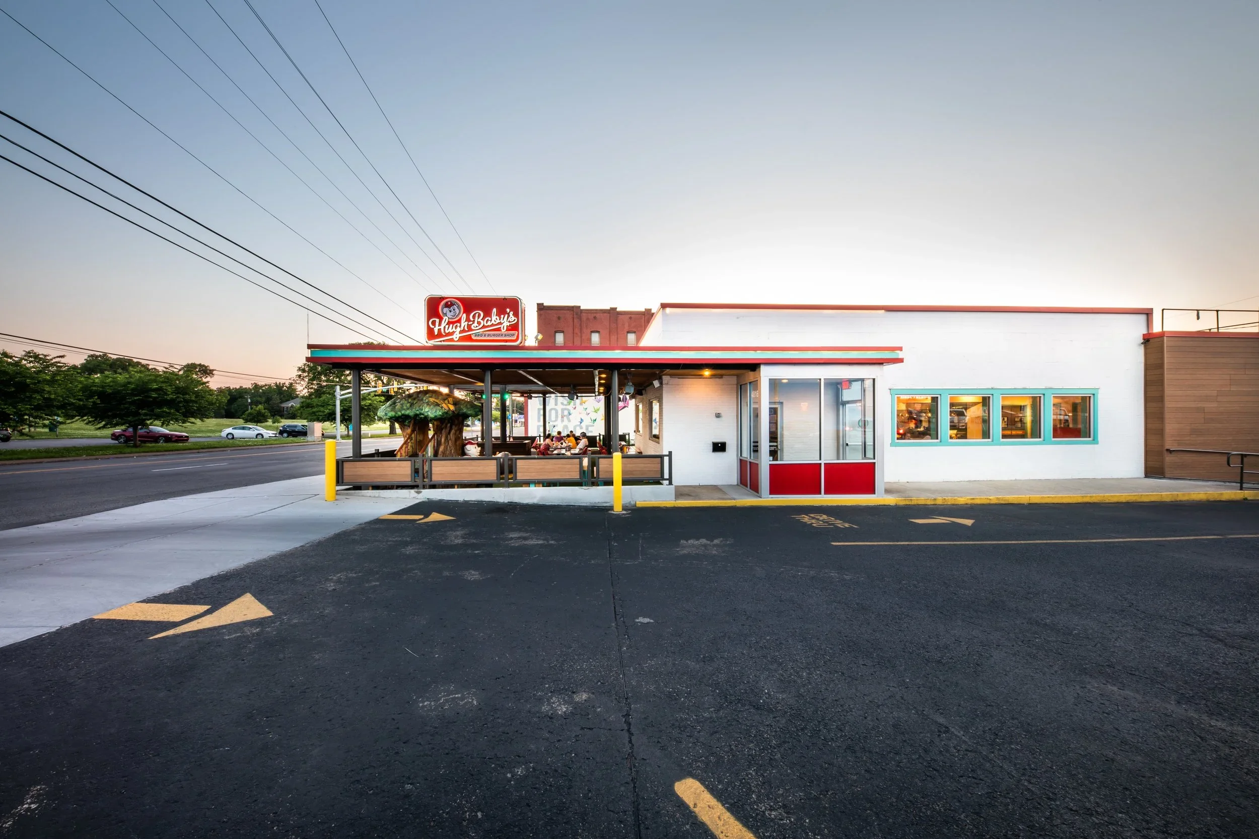 Fast food restaurant named Hugh Baby's BBQ & Burger Shop with outdoor seating, a neon sign, and bright red, white, teal, and wood accents, at dusk.