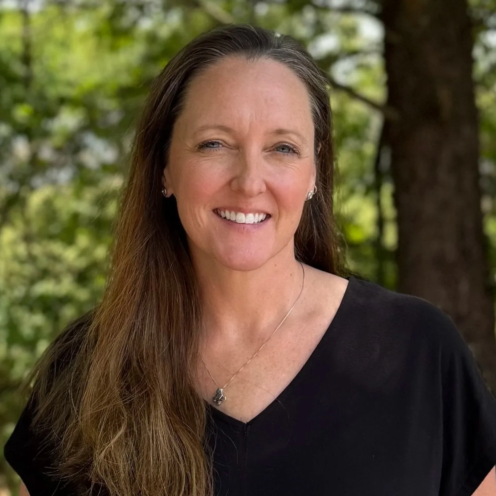 Close-up of a woman with long brown hair, smiling, wearing a black top, standing outdoors with trees and green foliage in the background.