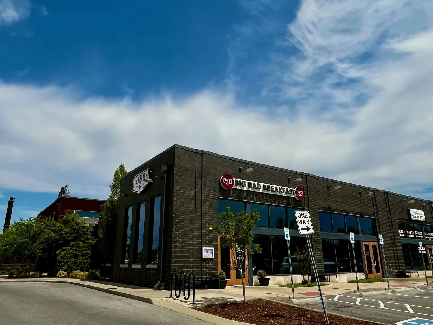 Exterior of a modern black brick building with a sign that reads 'Big Bad Breakfast,' large windows, trees, parking lot, and a blue sky with clouds.