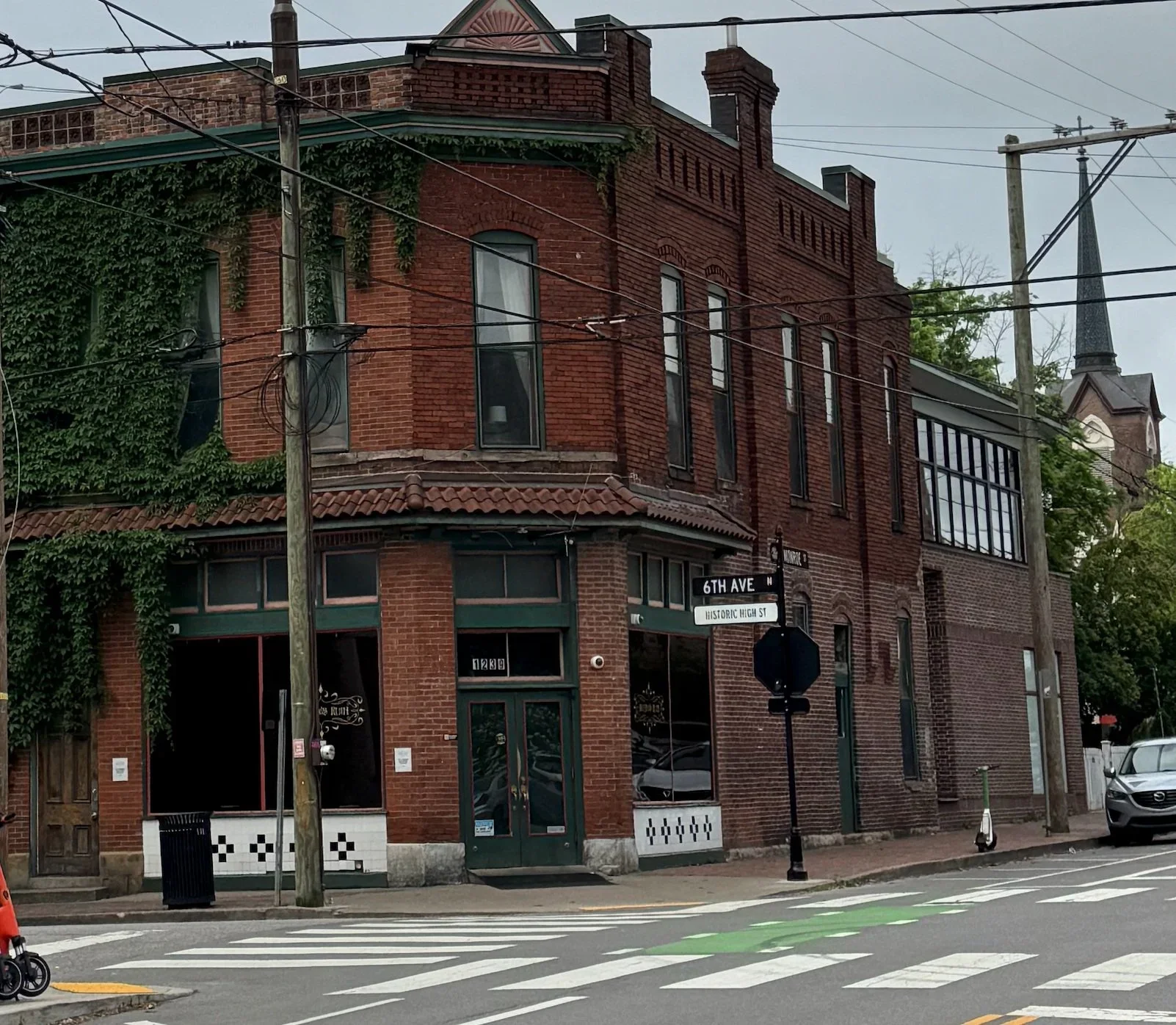 A historic brick building on a street corner with street signs for 6th Ave and Historic High St. The building has large windows, some with green frames, and ivy growing on parts of the facade. Utility poles and power lines are visible in front of the building, along with a crosswalk and a parked car.