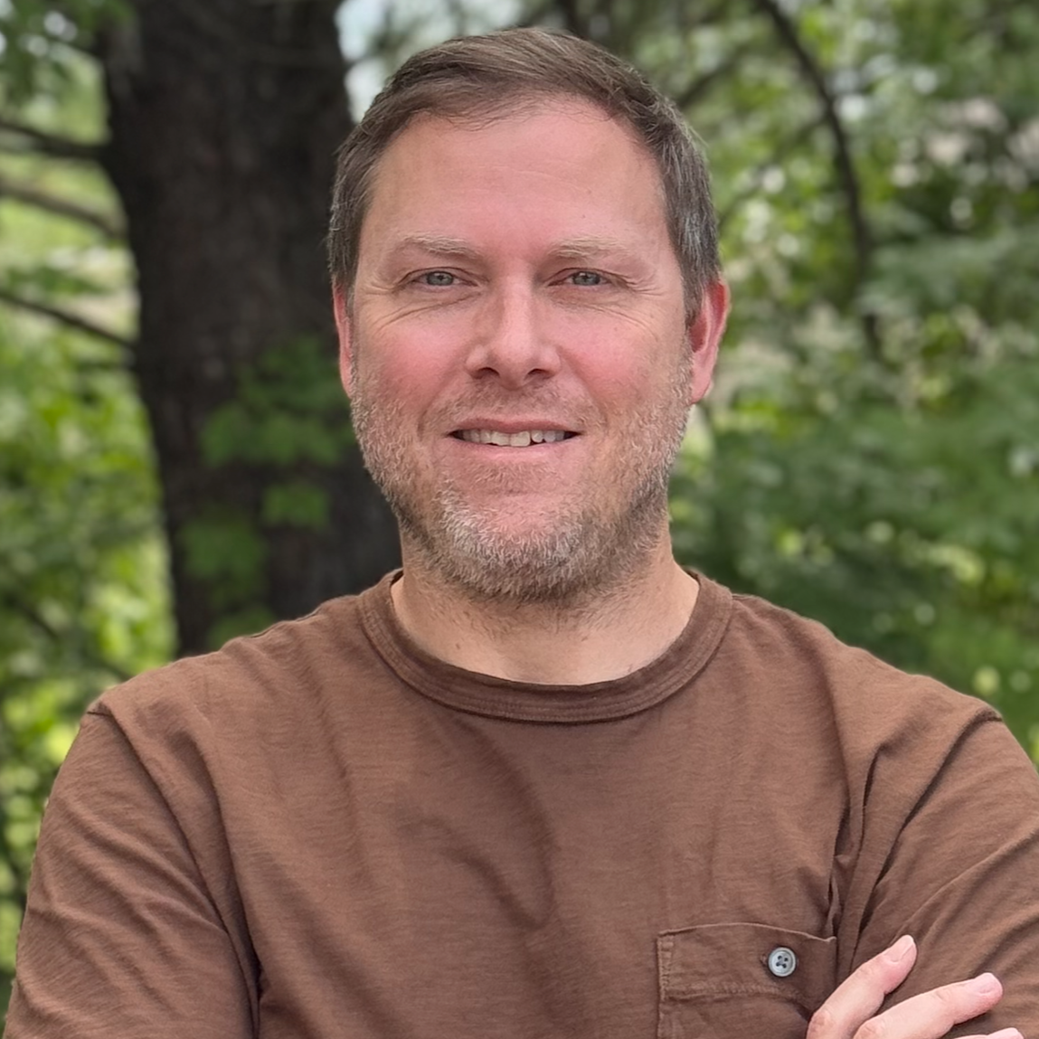 Man with short brown hair, light facial hair, wearing a brown shirt, smiling outdoors with greenery in the background.