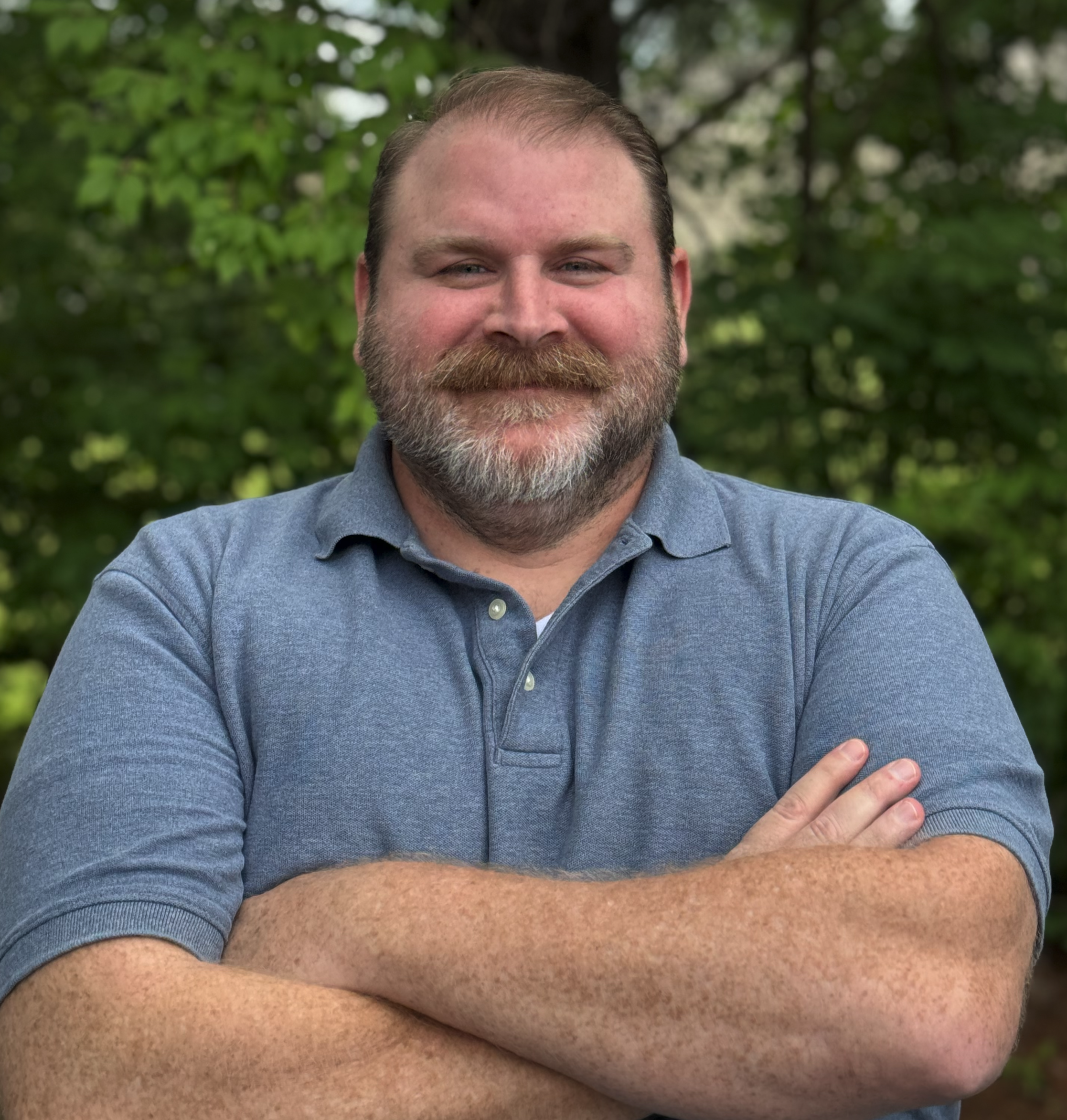 A man with a beard and mustache smiling outdoors among green trees, wearing a blue shirt.