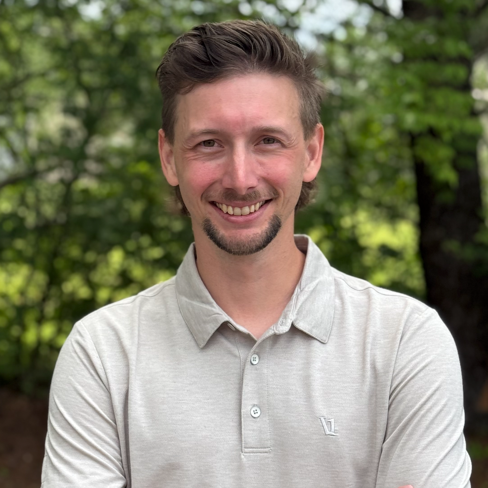 A smiling man with short brown hair and facial hair standing outdoors with a green leafy background.