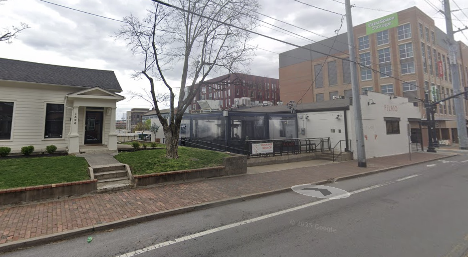 Street view showing a small white house with a black door on the left, a leafless tree in front, and a larger brick building with green sign that says ExtraSpace Storage on the right. The sky is cloudy.