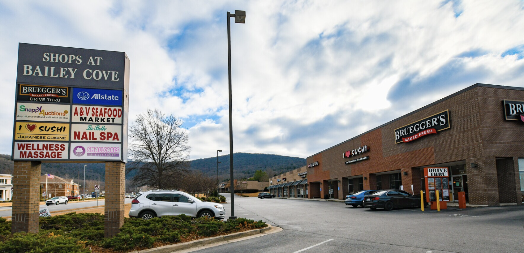 Parking lot in front of Bailey Cove shopping center with signs advertising shops including Bruegger's Baked Fresh, Allstate, A & V Seafood Market, Japanese Cuisine, Nail Spa, Wellness Massage, Smallcakes, and an All You Auctions drop-off site. There are several cars parked, a brick building with store signs, a blue sky with clouds, and hills in the background.