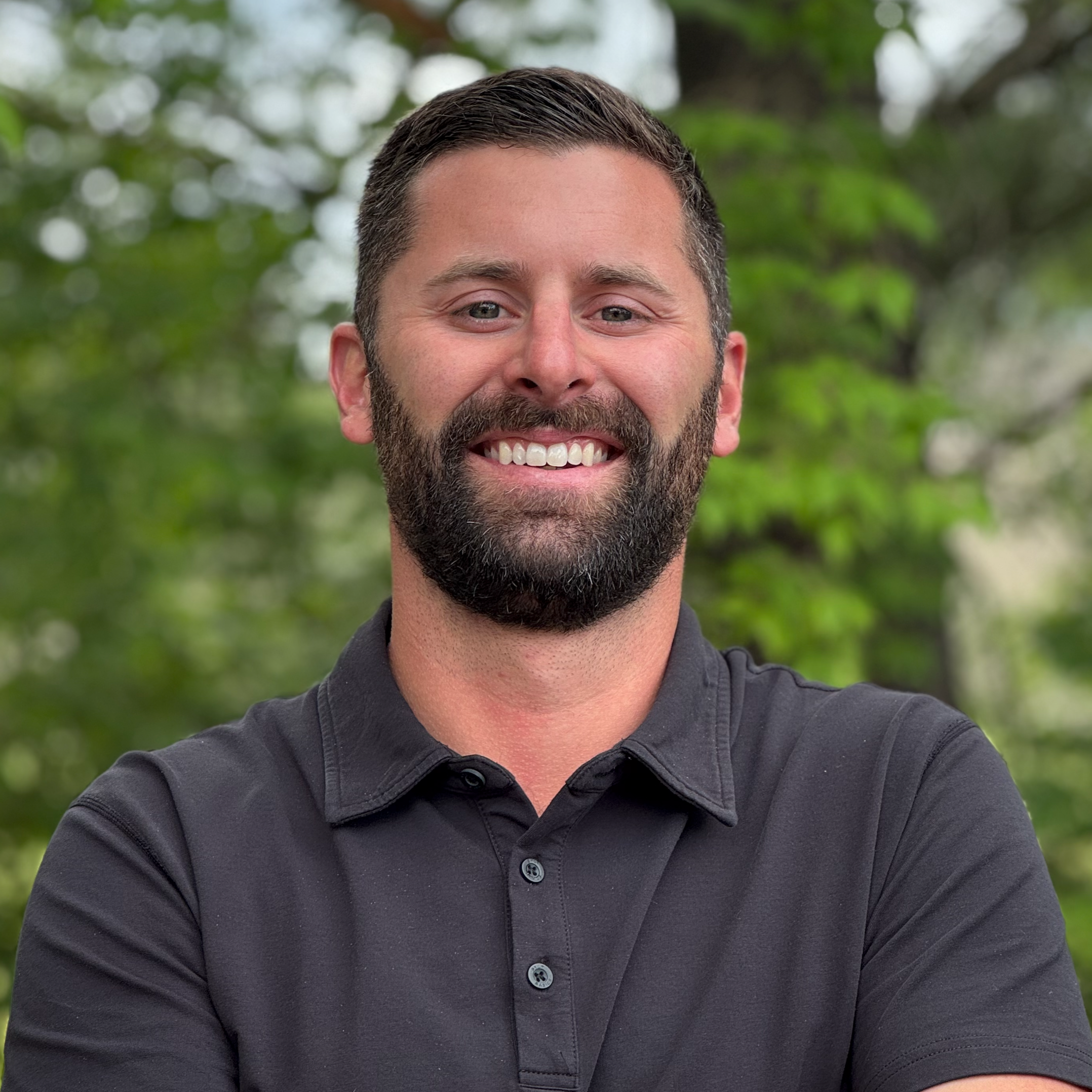 A man with dark hair and a beard smiling, wearing a dark gray collared shirt, with a blurred green outdoor background.