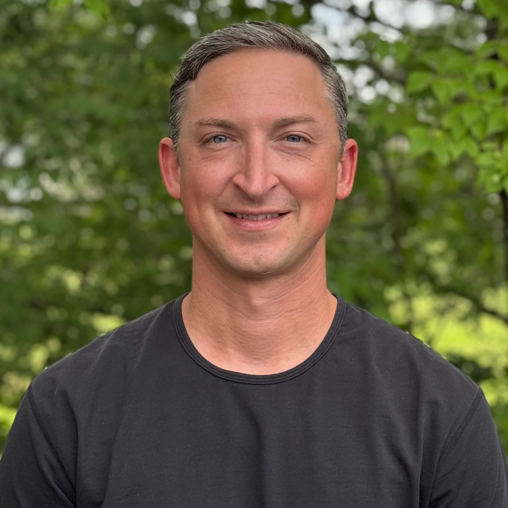 A smiling man with short, dark gray hair, wearing a black T-shirt, standing outdoors with a background of lush green trees.