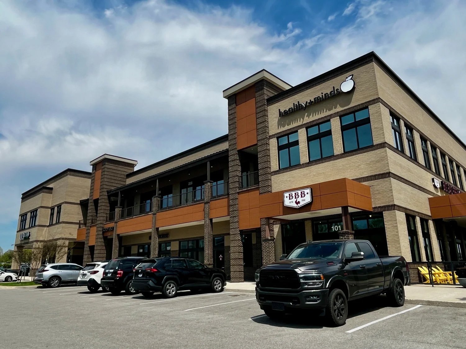 A modern two-story shopping center with parked cars in front. The store signs include 'healthy + minds' and 'BBB.' The building has brick and beige siding with orange accents. The sky is partly cloudy.