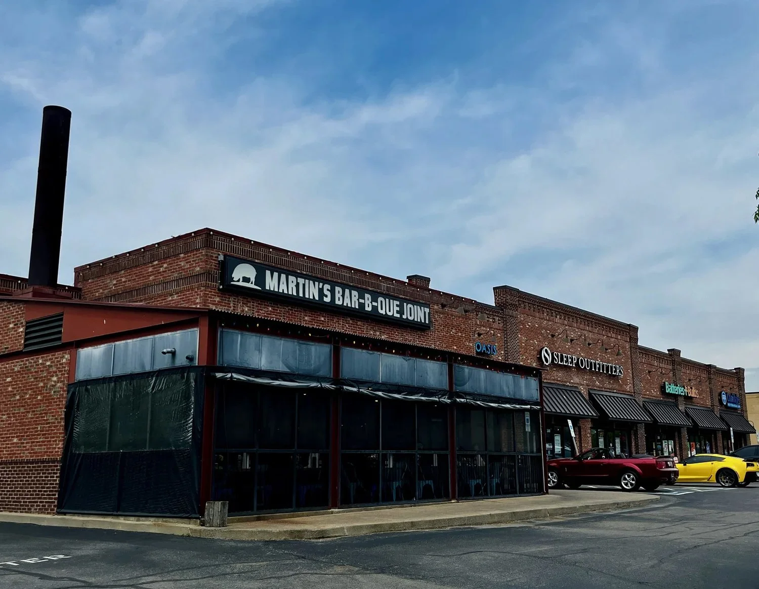A strip mall made of red brick with signs for various stores including Martin's Bar-B-Que Joint, Sleep Outfitters, Oasis, Batteries Plus, and a blue sign with Frito Lay logo. There are a few parked cars in front, and the sky is partly cloudy.