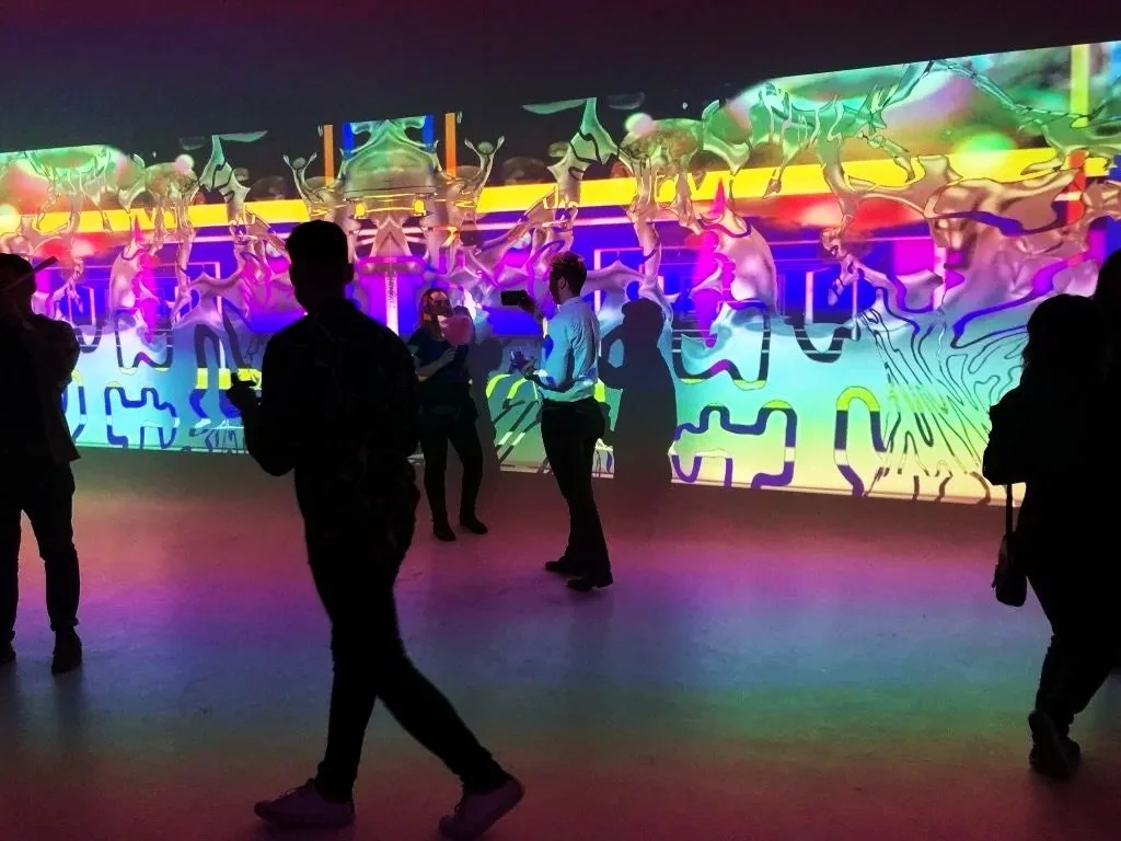 People standing and walking in a dark room with a colorful, abstract, immersive, illuminated digital art display on the wall.