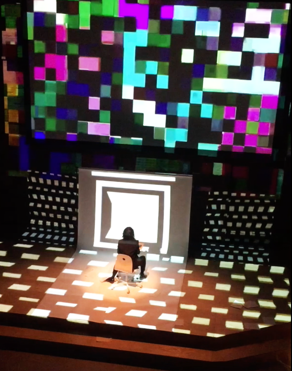 Sook YIn Lee sitting on an office chair on stage with a bright lighted square behind them, surrounded by a digital pixelated display with colorful squares and abstract shapes projection mapped on stage.