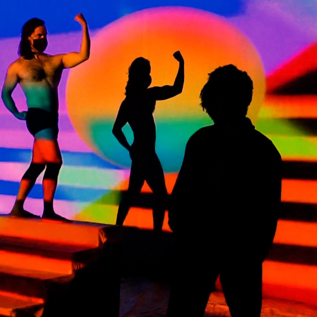 Silhouettes of three people, two women and one man, posing with flexed arms against a vibrant, colorful sunset background projected on a screen.