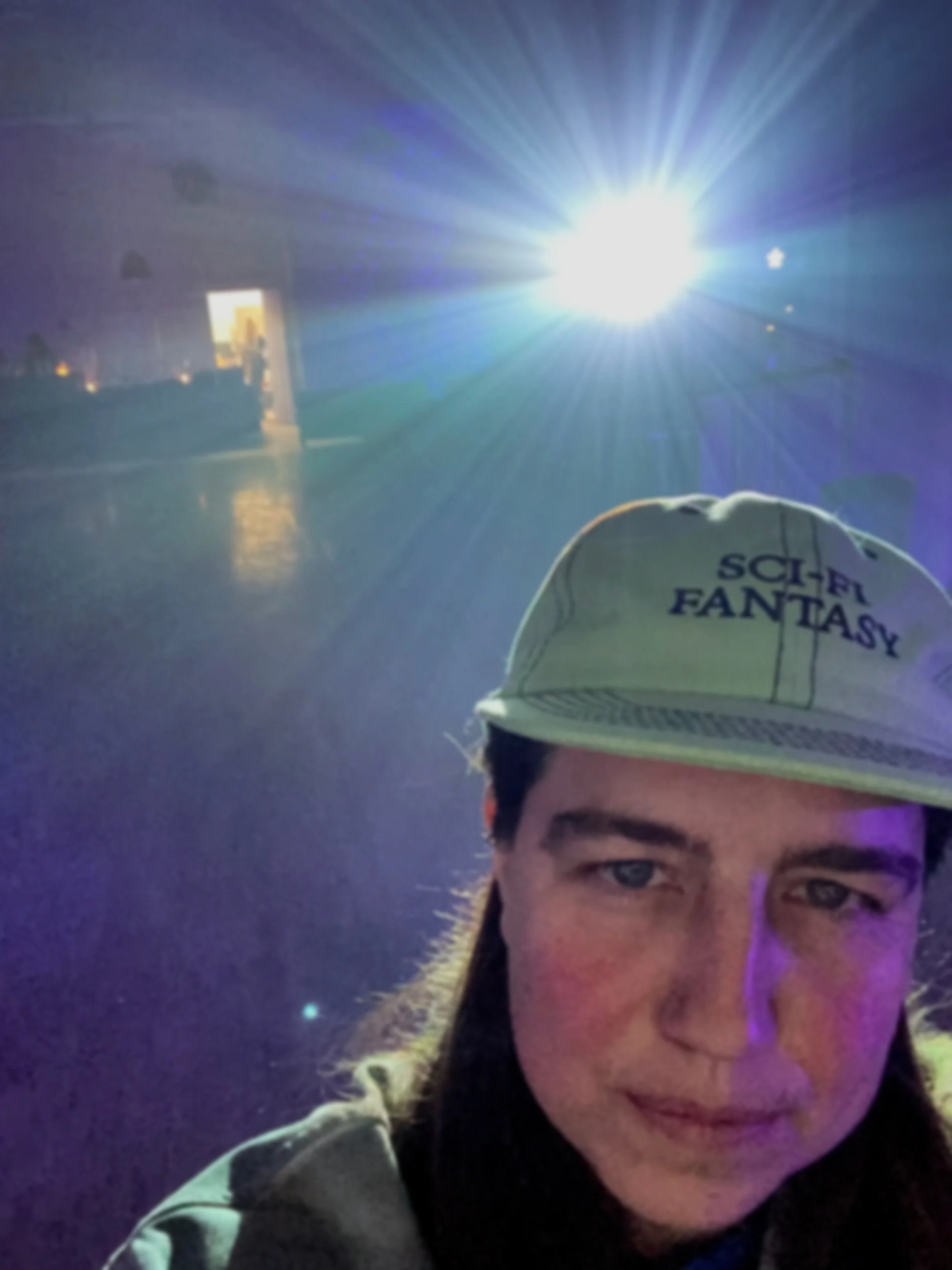 A person taking a selfie in front of a bright light with purple and blue hues. The person is wearing a white cap with the words 'SCI-FI FANTASY' and has long brown hair and a slight smile.