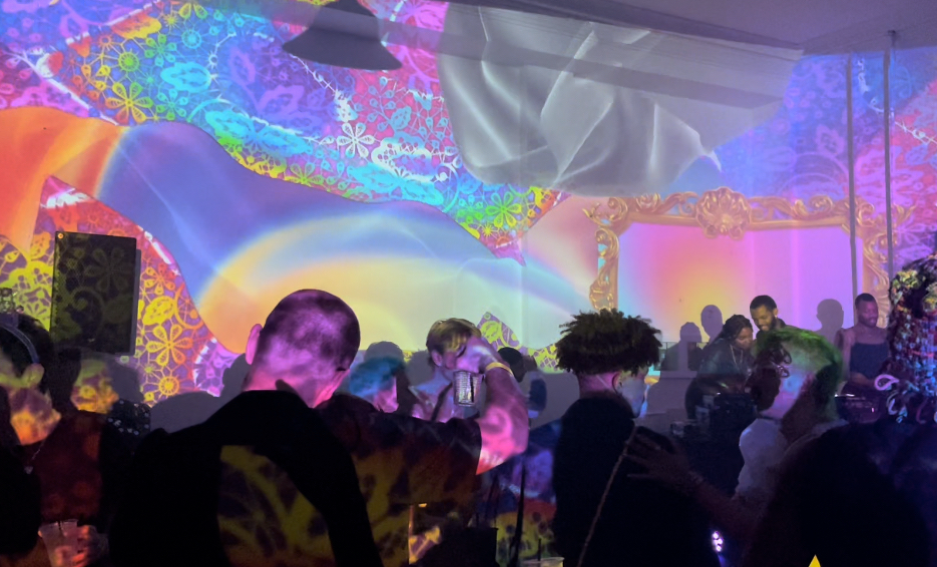 People dancing and socializing at a party with colorful, vibrant light projections and decorations in the background.