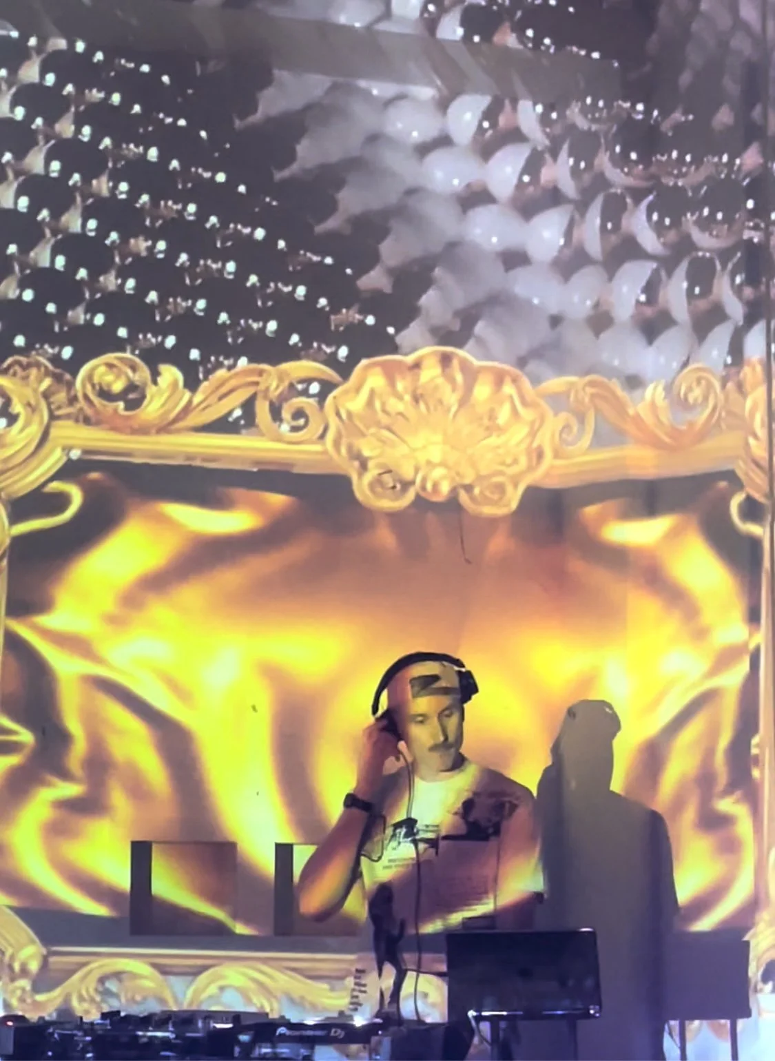 A DJ wearing headphones controls music at a concert with a yellow and orange fire-themed digital backdrop.