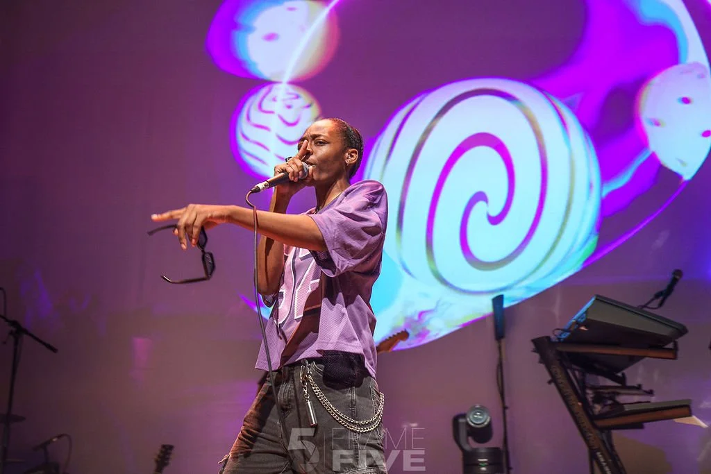A performer singing into a microphone on stage with colorful abstract art projected in the background.
