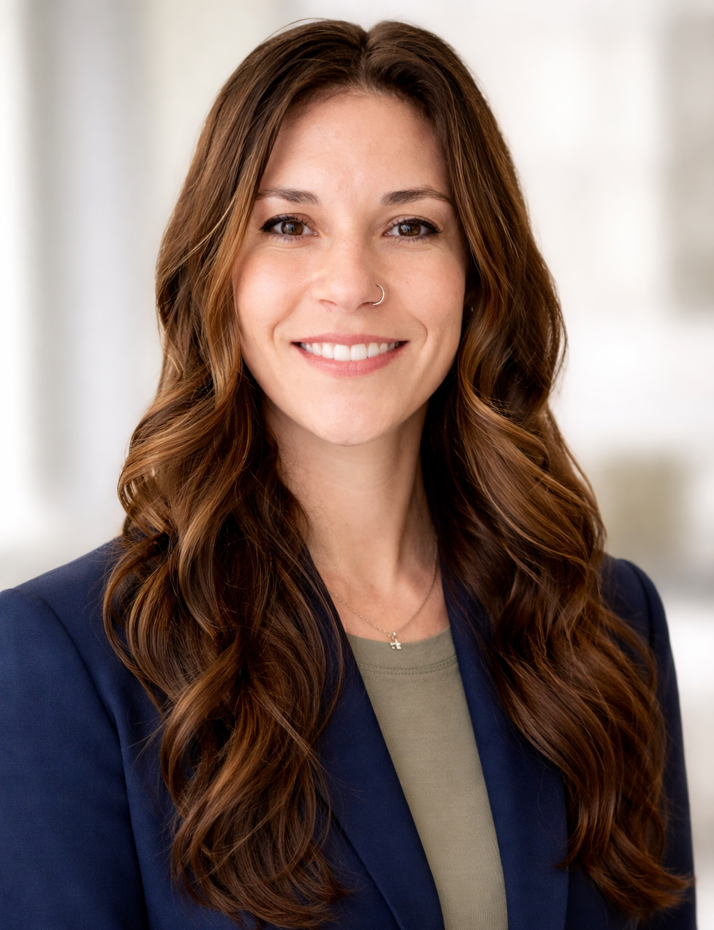 A woman with long, wavy brown hair and a nose ring, smiling while dressed in a navy blazer and a light green top, in a professional setting.