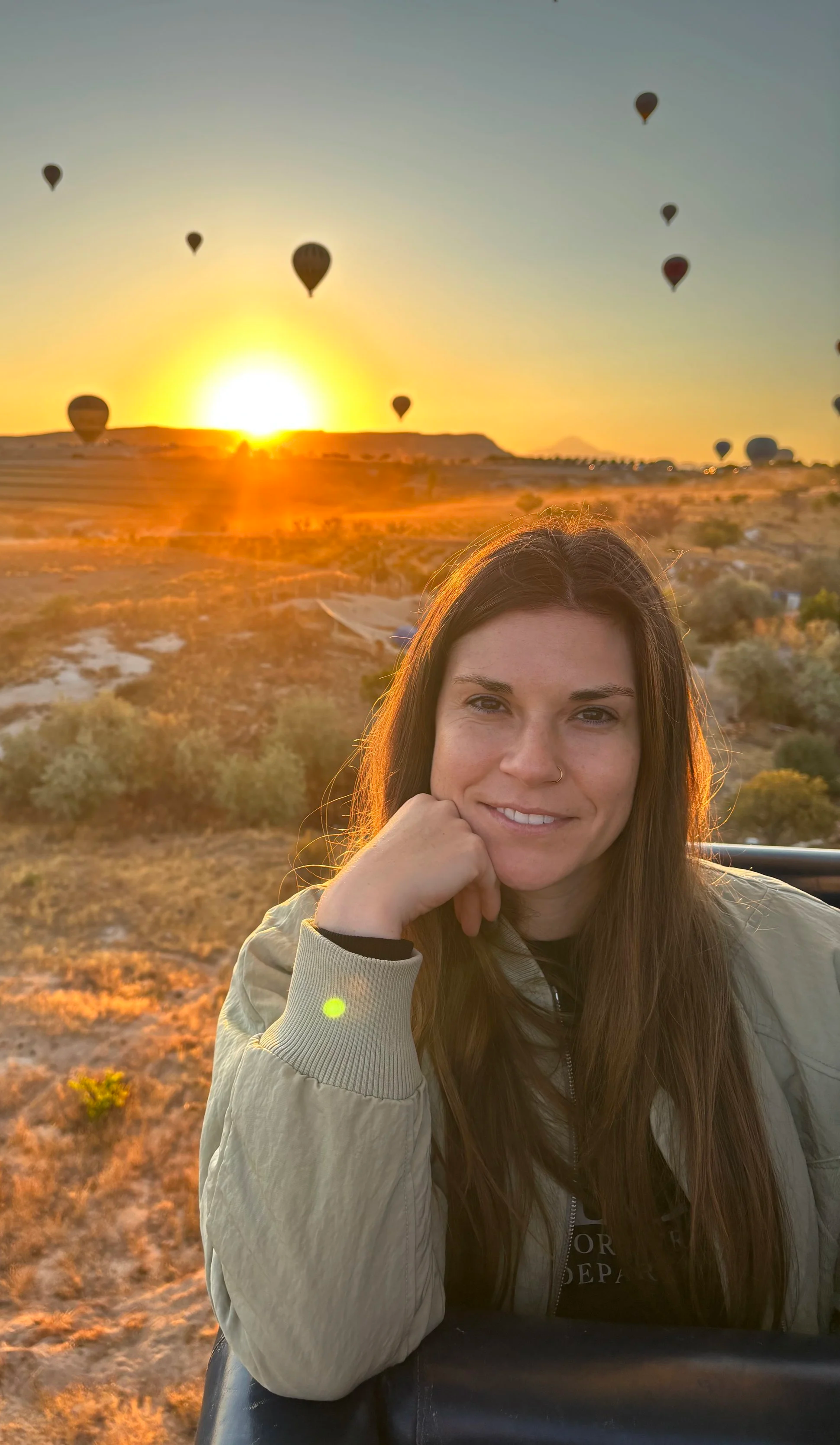 A woman with long brown hair, smiling, resting her chin on her hand, in a beige jacket, during a sunset in a hot air balloon with other balloons floating in the sky over a desert landscape.