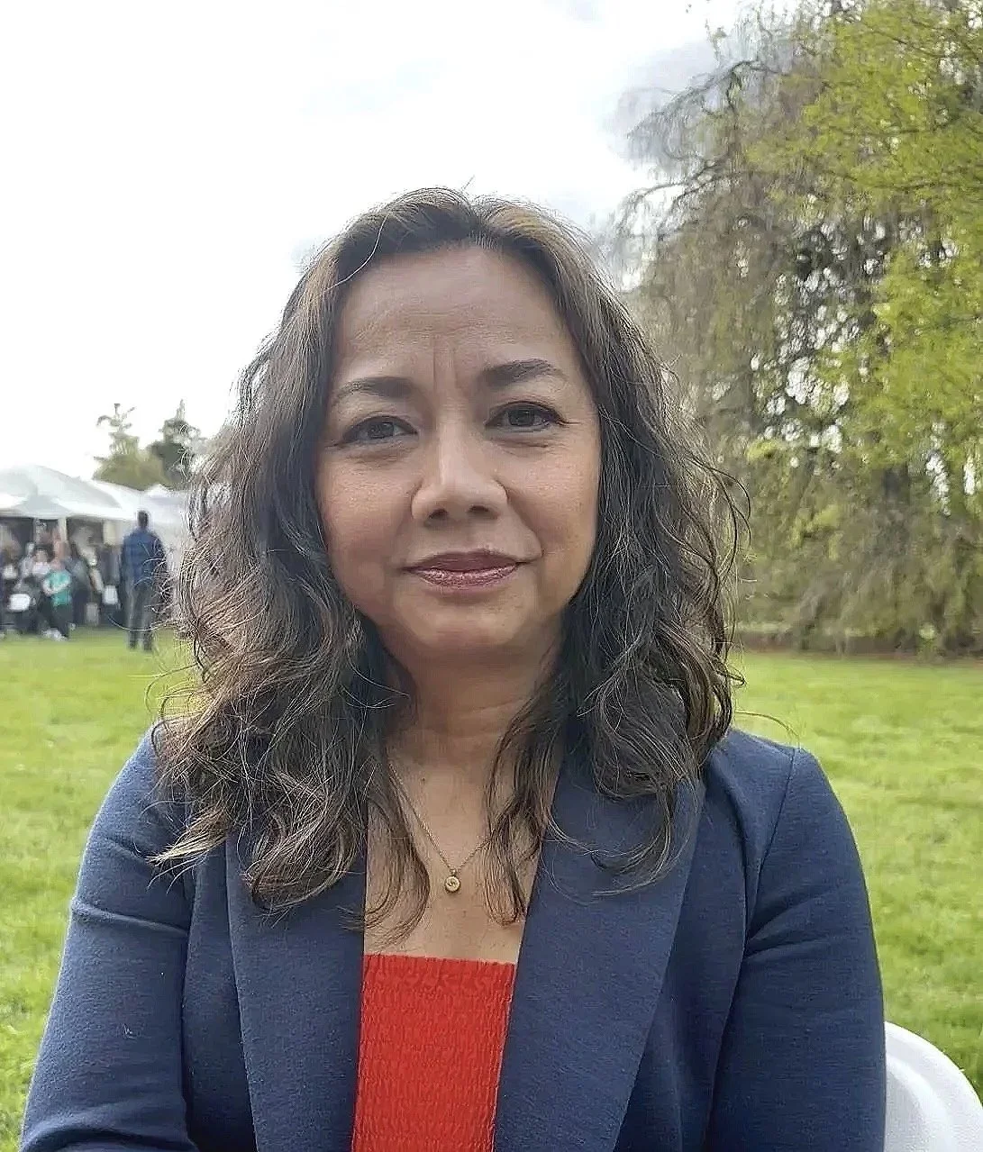Chulee, a woman with dark wavy hair wearing a blue blazer and red top, sitting outdoors on a green lawn with trees and a cloudy sky in the background.
