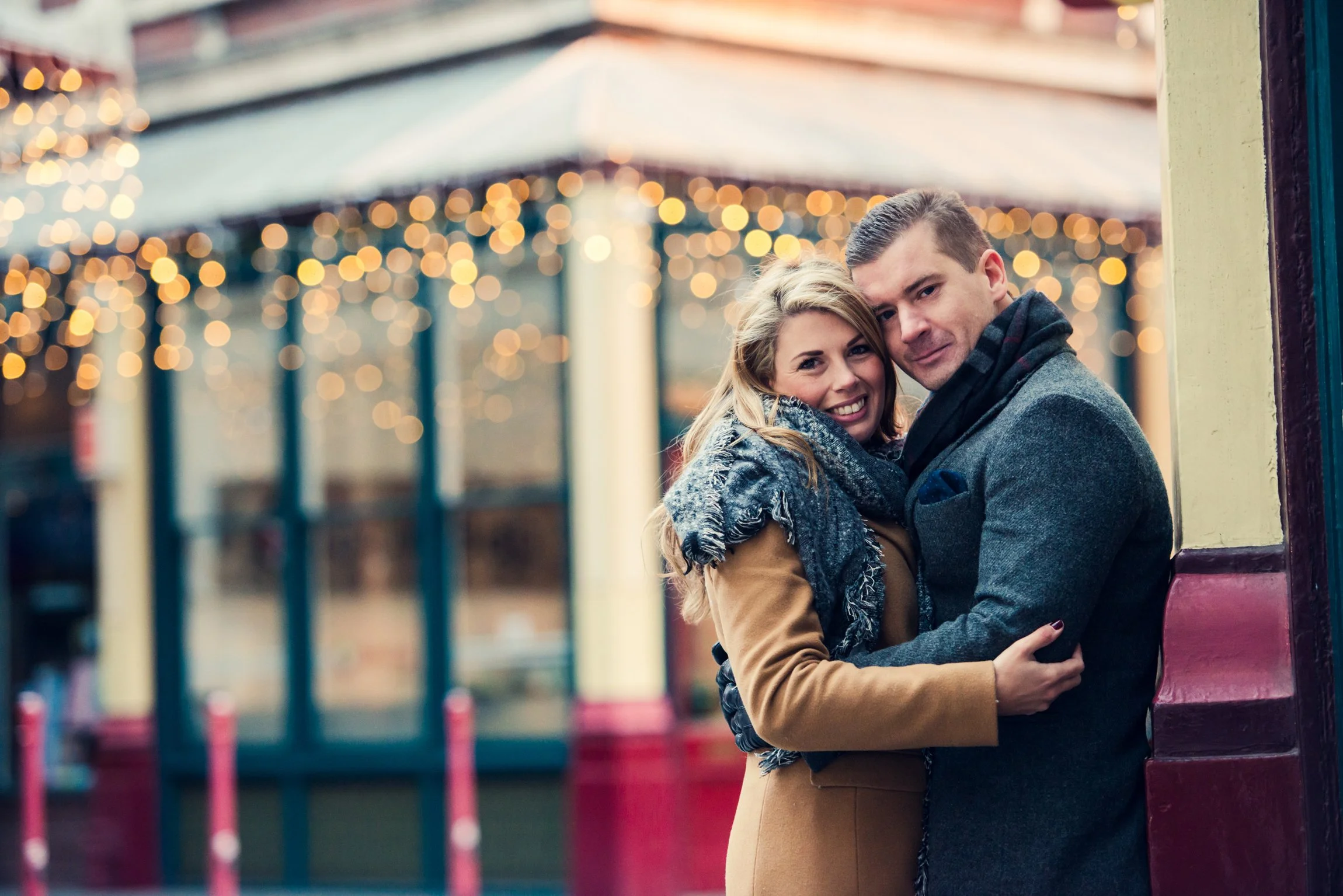 A smiling couple is standing close together outside near a building with colorful trim. The woman is wearing a tan coat and a blue scarf, while the man is dressed in a gray coat and dark scarf. In the background, festive string lights are blurred, creating a warm, bokeh effect.