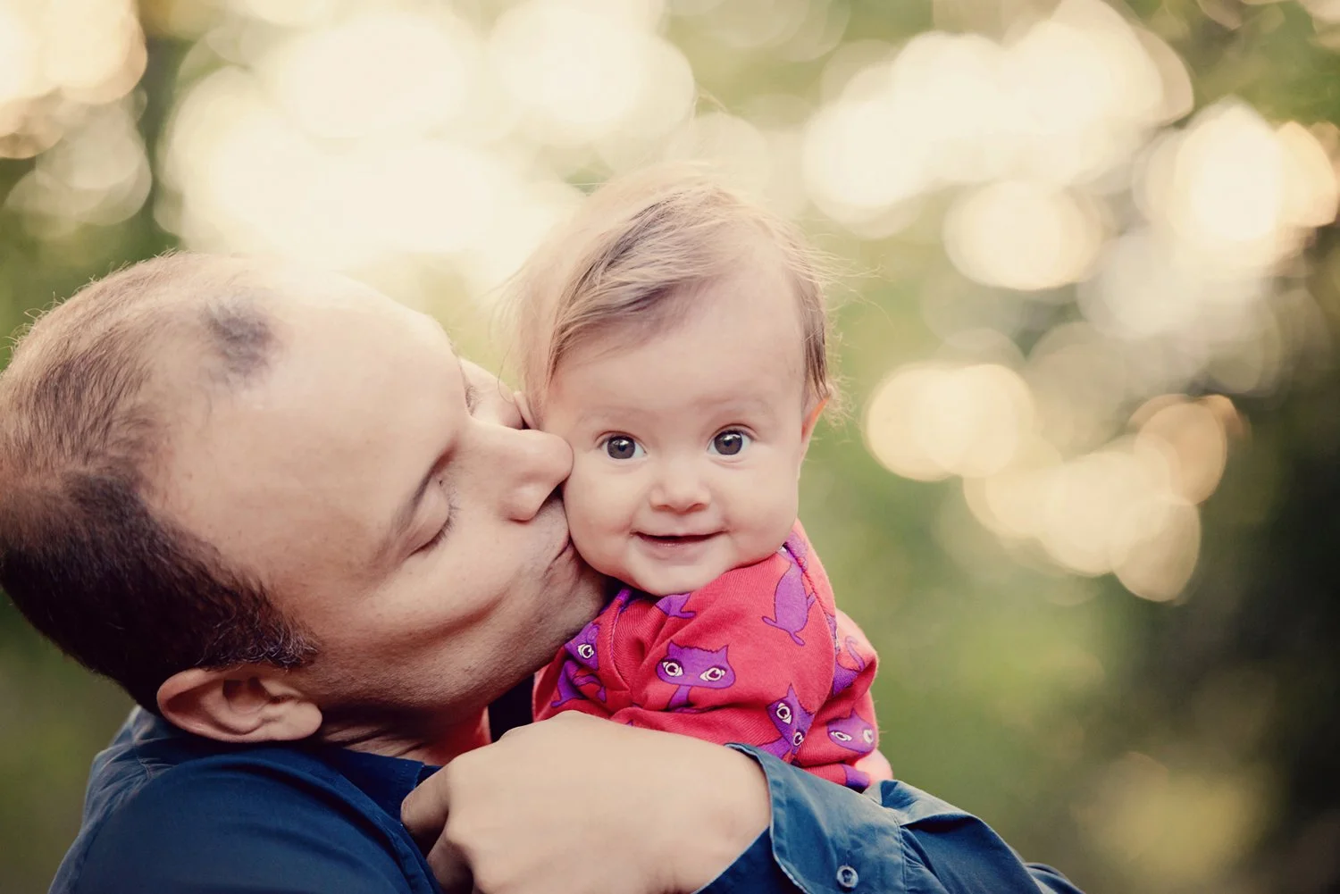 A man kissing a baby girl on the cheek outdoors with blurred foliage in the background.
