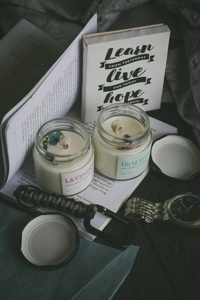 Open book with motivational quotes, two scented candles in glass jars labeled 'Lavender' and 'Fiji Breeze,' a wristwatch, and a curling iron on a dark wooden surface.