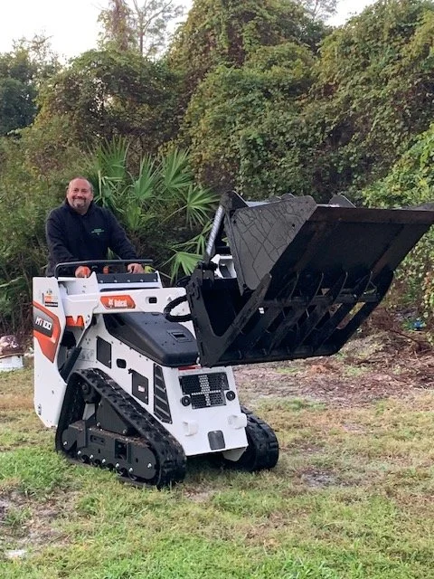 A man smiling and sitting in a small white and black excavator with a raised bucket, outdoors on grass with trees in the background.