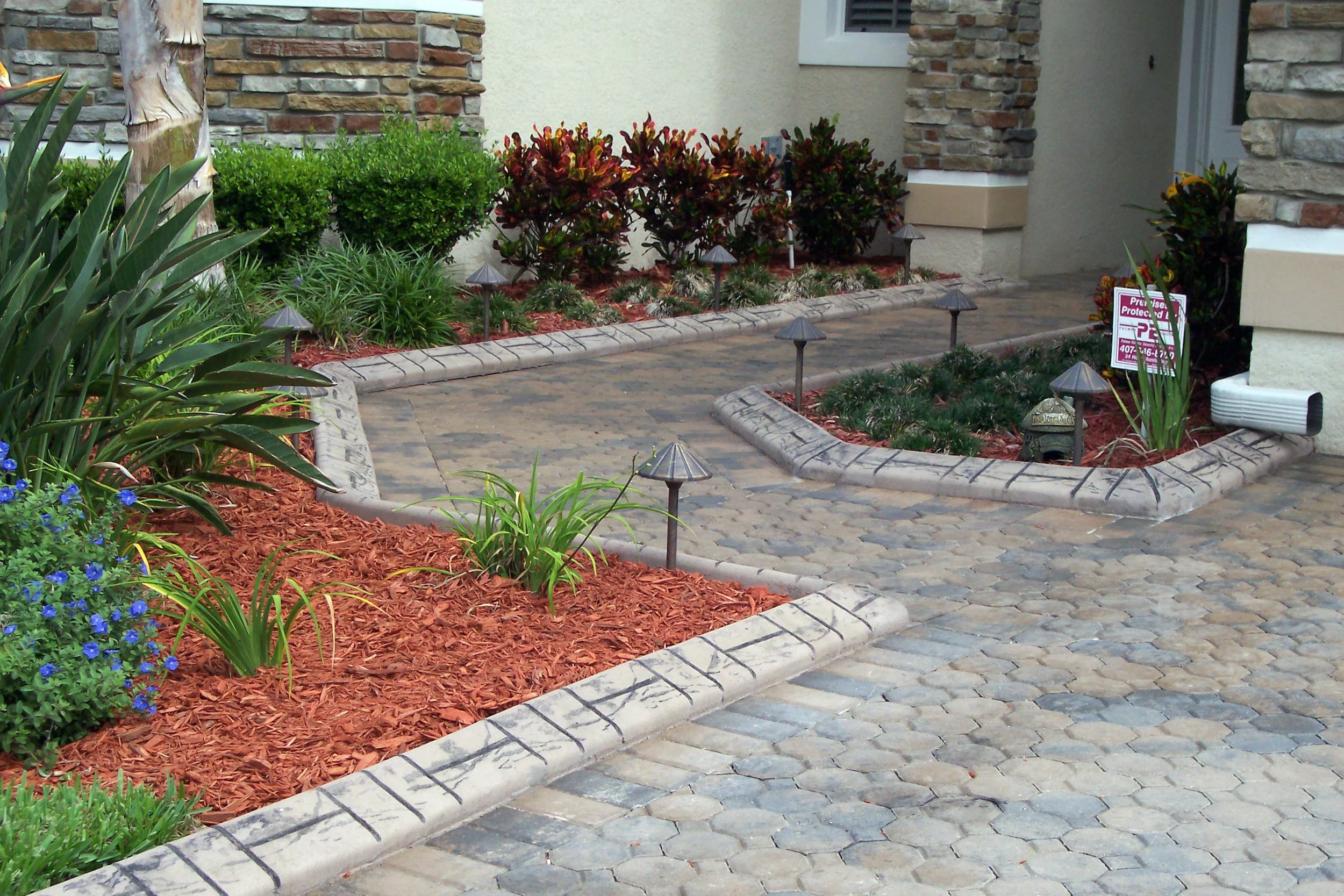 Paved driveway and walkway with landscaped garden beds containing plants and mulch, surrounded by brick edging, with a house wall in the background.