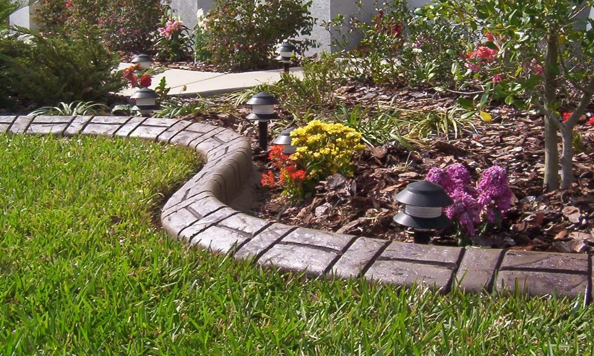 Flower bed with colorful flowers and small pathway, surrounded by grass, with landscape lights illuminating the area.