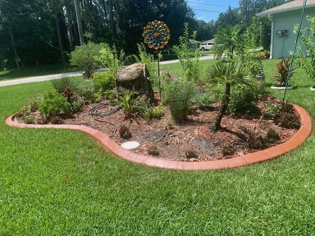 A small garden bed with various plants, a large rock, and a decorative wind spinner in a residential yard, bordered by red bricks.