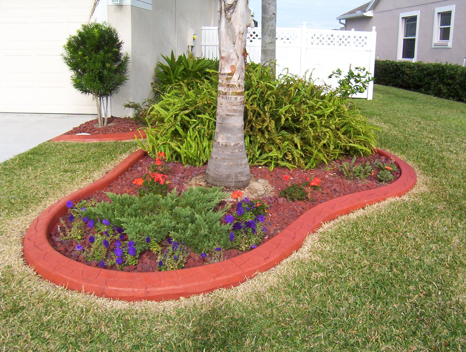A landscaped yard with a brick-edged flower bed containing a small palm tree, various green plants, and colorful flowers, surrounded by well-manicured grass and a white fence.
