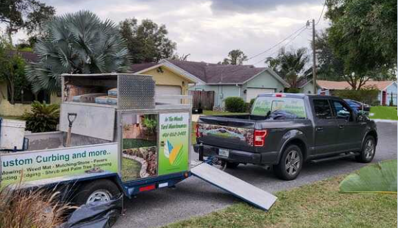 A small mobile tree trimming and debris removal service trailer attached to a black pickup truck parked on a residential street with trees and houses in the background.