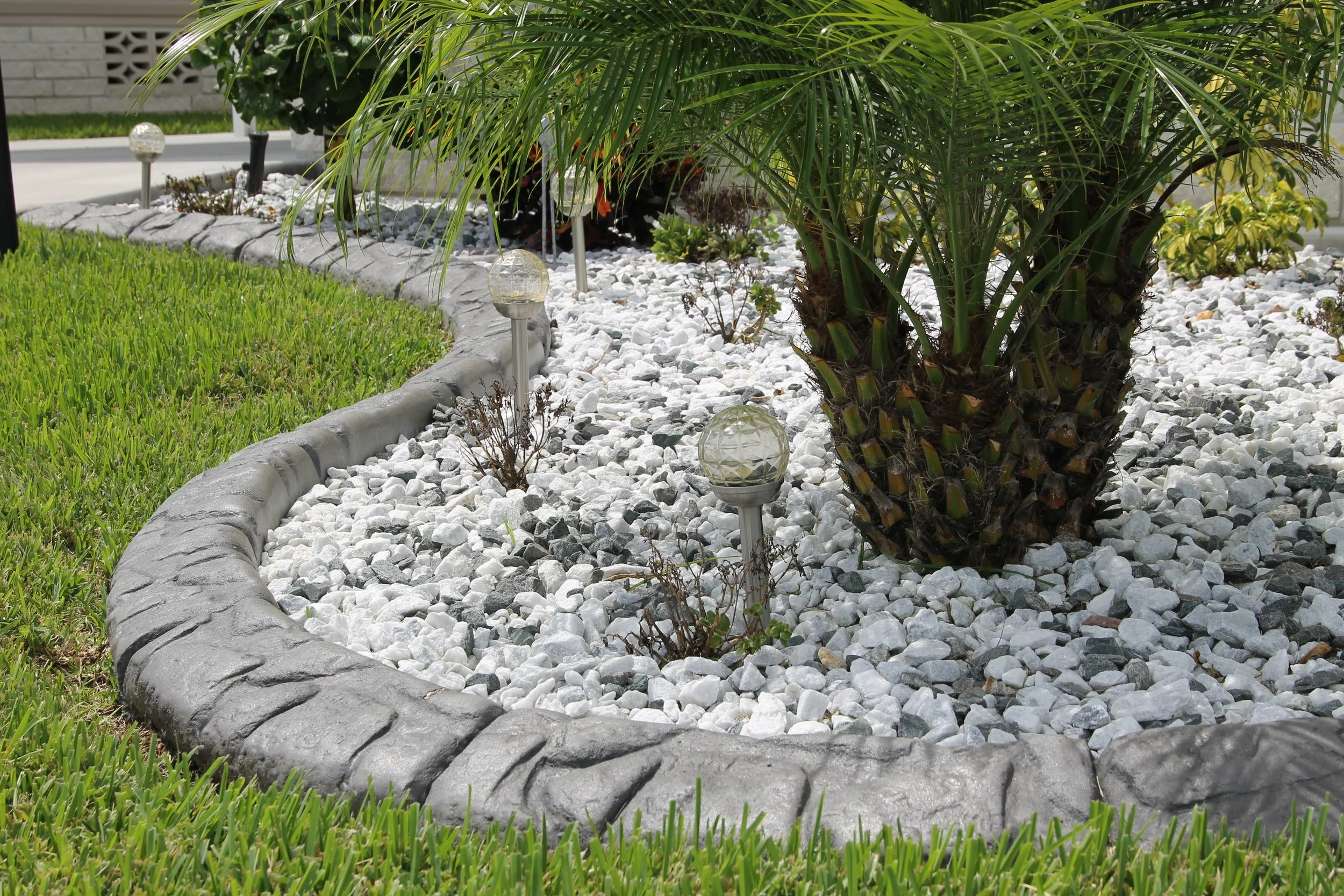A landscaped yard with a curved stone border, green grass, white decorative gravel, small plants, and a palm tree, illuminated by solar pathway lights.