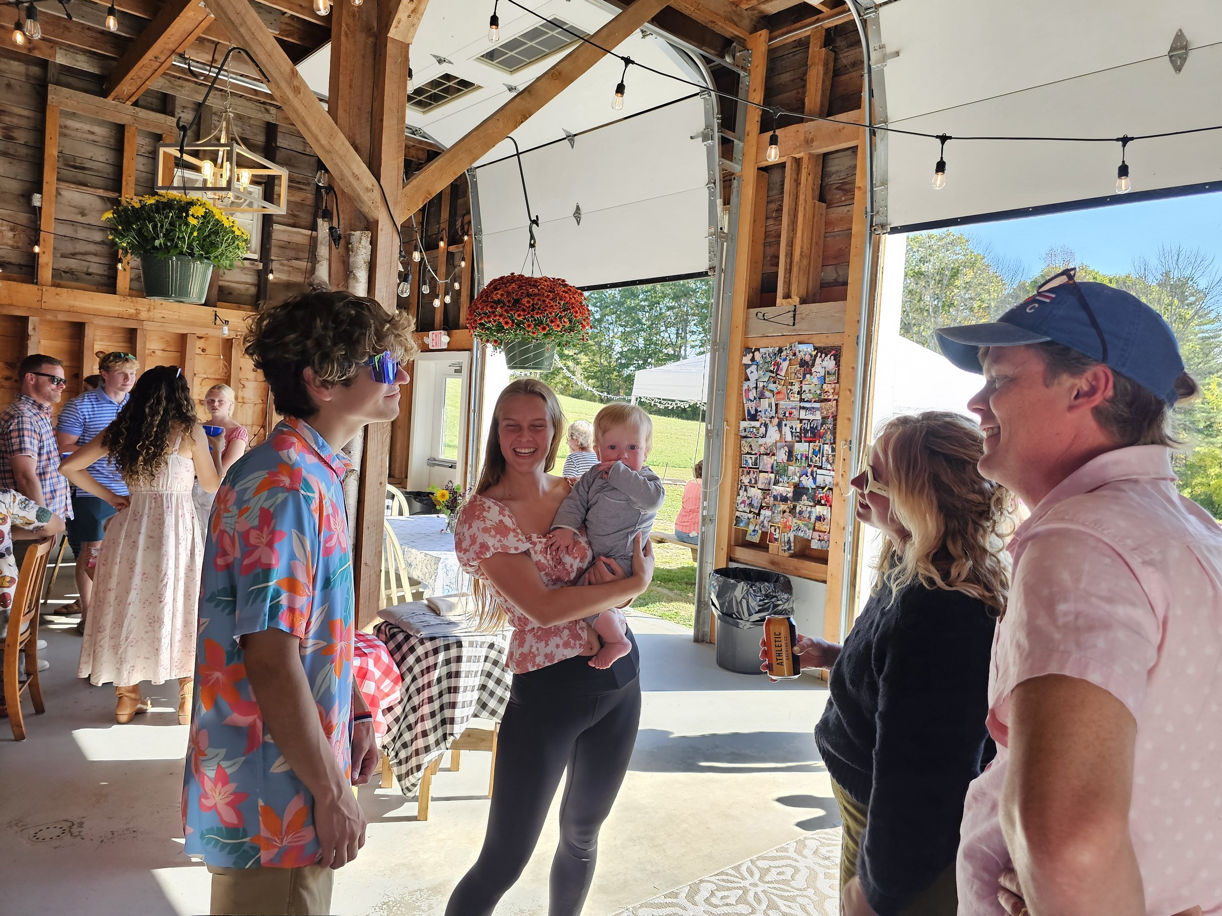 Group of people at an outdoor gathering socializing inside a wooden barn decorated with hanging string lights, flower pots, and a collage of photos on the wall, with some individuals smiling and talking.