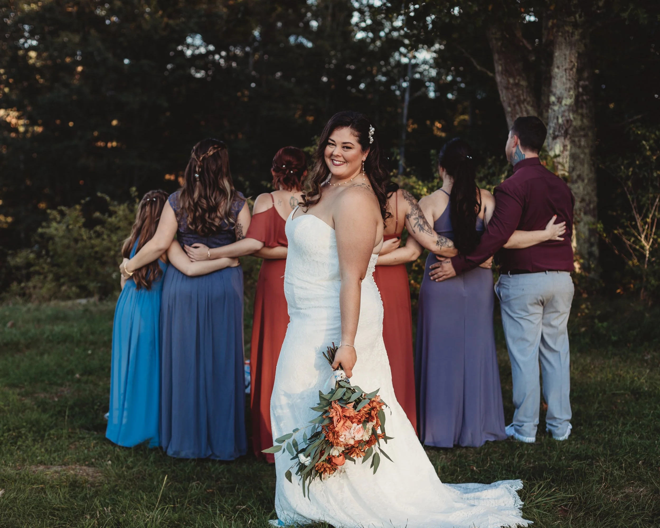 Bride in a white wedding dress holding a bouquet of orange and green flowers, smiling at the camera, while her friends stand in a semi-circle with their backs to her, outdoors with trees in the background.