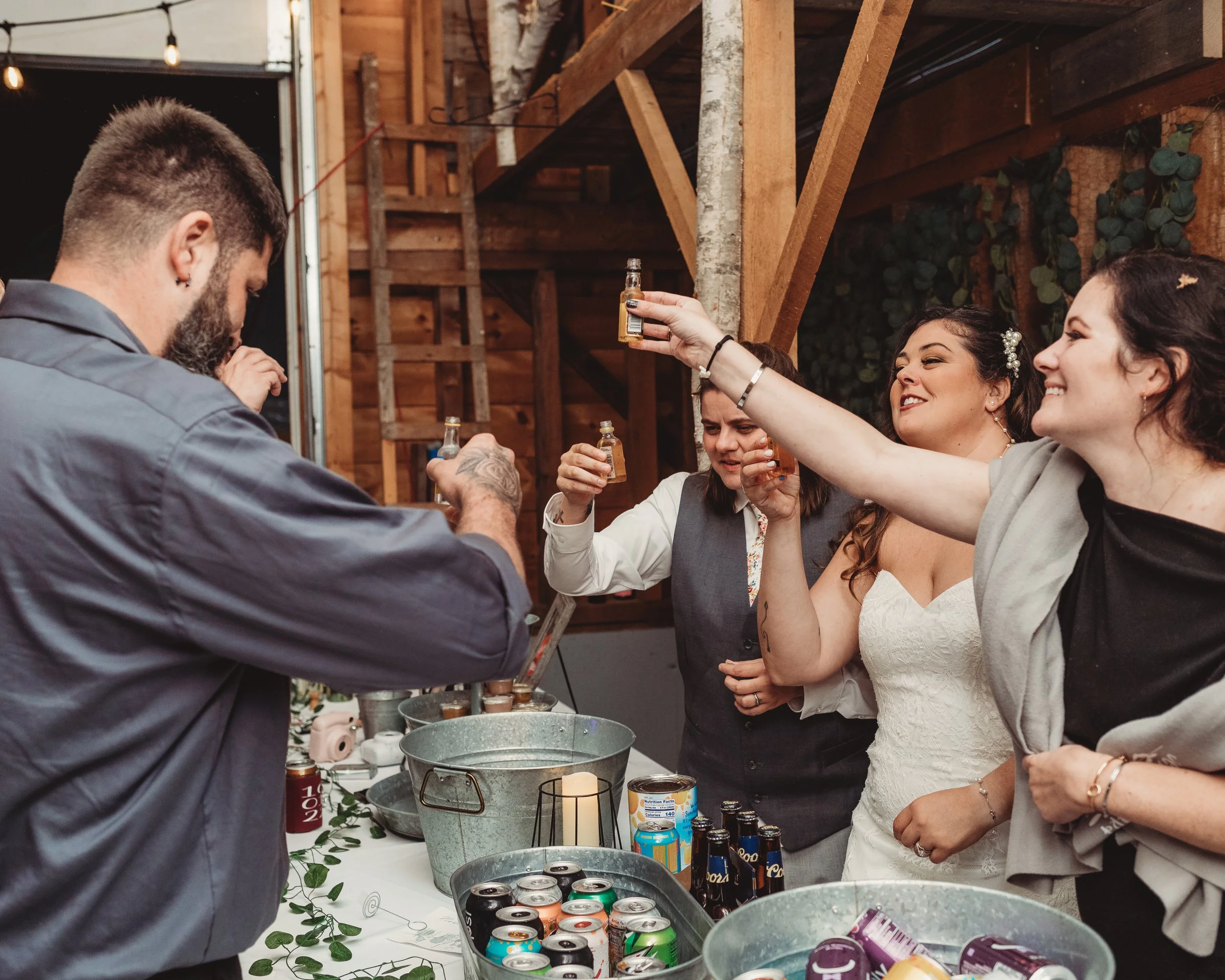 Group of friends toasting with drinks at a party in a rustic wooden venue, with a table of beverages and snacks.