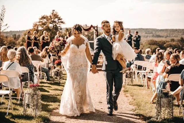 A newlywed couple walking down an outdoor aisle during a wedding ceremony, with guests seated on either side and a young girl in a white dress sitting on the groom's arm.