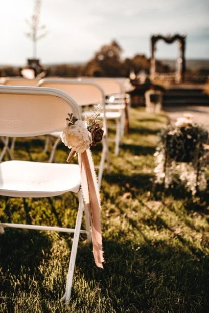 Setting for an outdoor wedding ceremony with white chairs decorated with flowers and ribbons, and a wedding arch in the background on a grassy area.