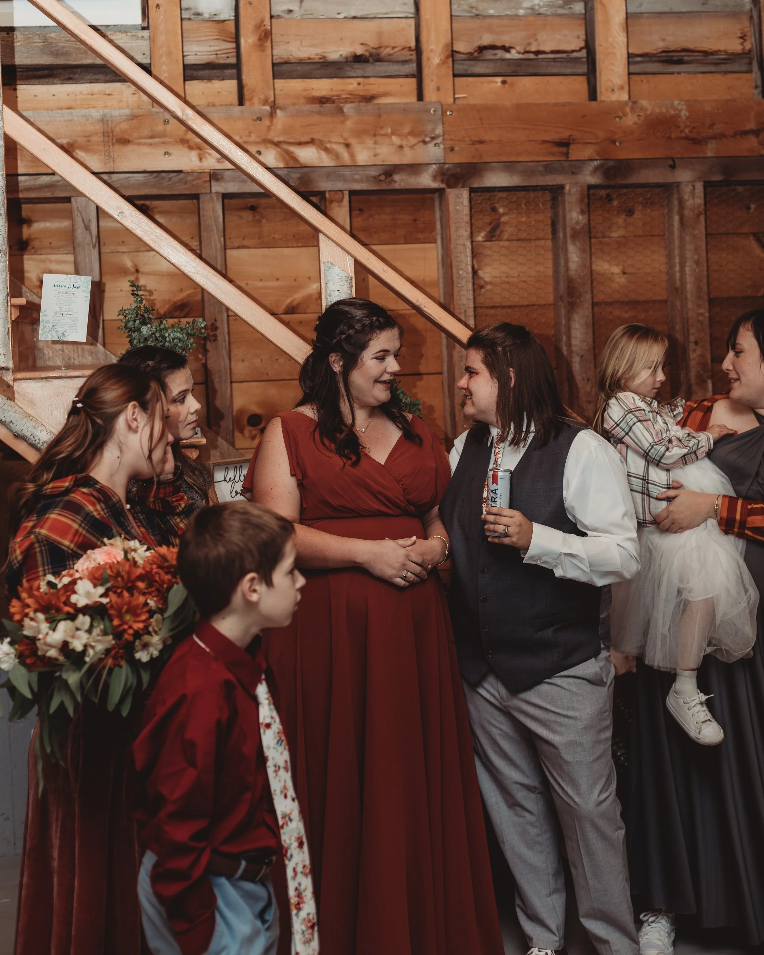 Group of people, mostly women, socializing at an indoor event with wooden walls. One woman is holding a bouquet of flowers, and a young girl is being held by an adult in the background.