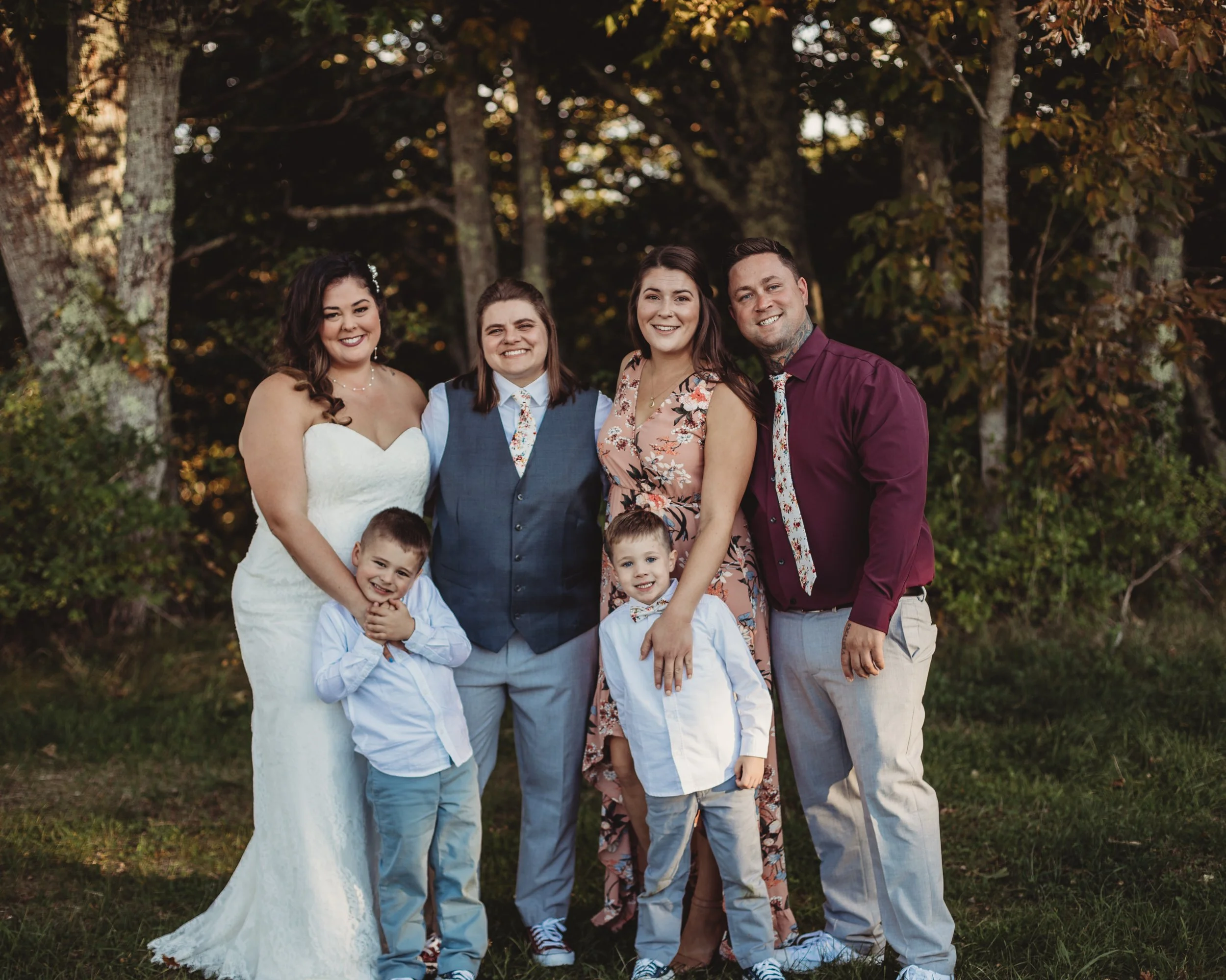 A family of six posing outdoors in front of trees in the evening. The mother is wearing a white wedding dress, the father in a maroon shirt, and children dressed in light-colored clothes. The group is smiling and standing on grass.