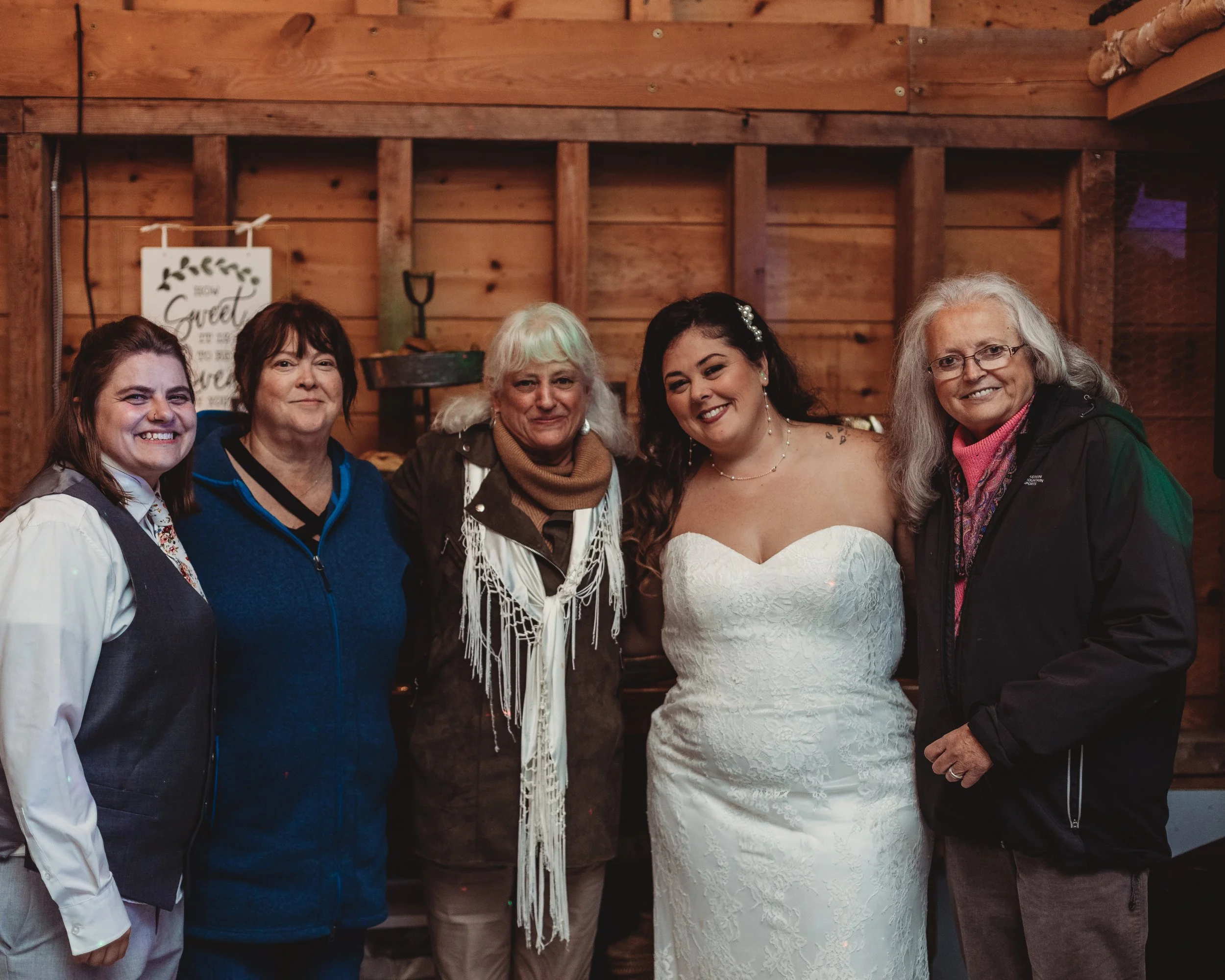A group of five women standing together indoors, four of them dressed casually and one in a wedding gown, smiling at the camera.