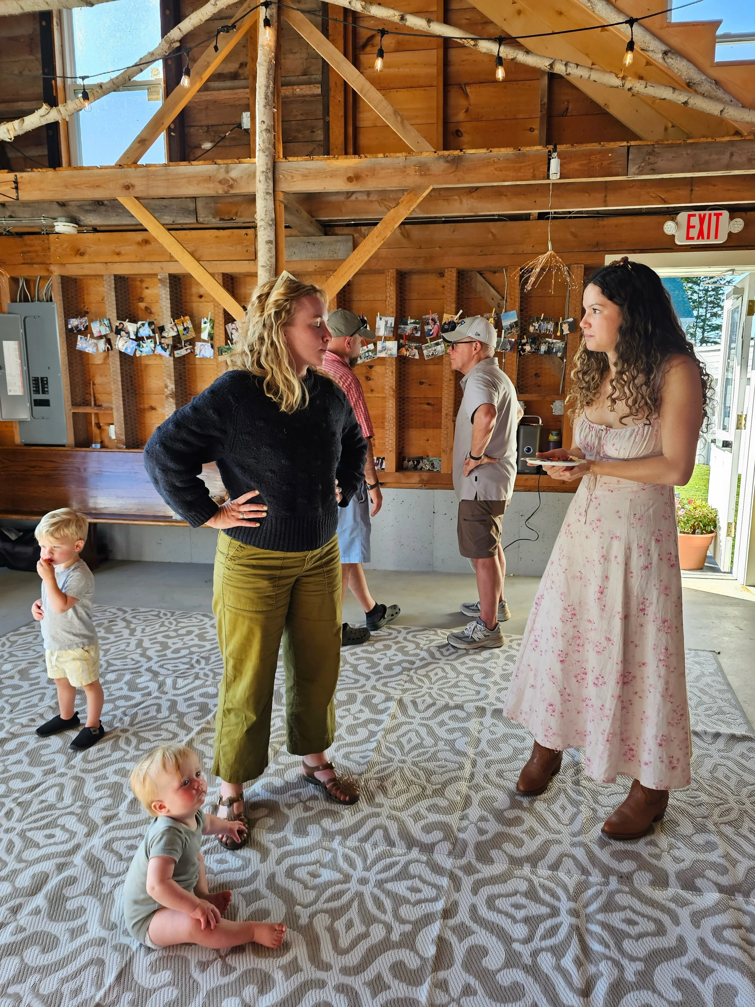 Indoor gathering with five adults and three children. Two women are engaged in conversation, one in a black sweater and olive green pants, the other in a floral dress. Two men are in the background, one in a plaid shirt and baseball cap, the other in