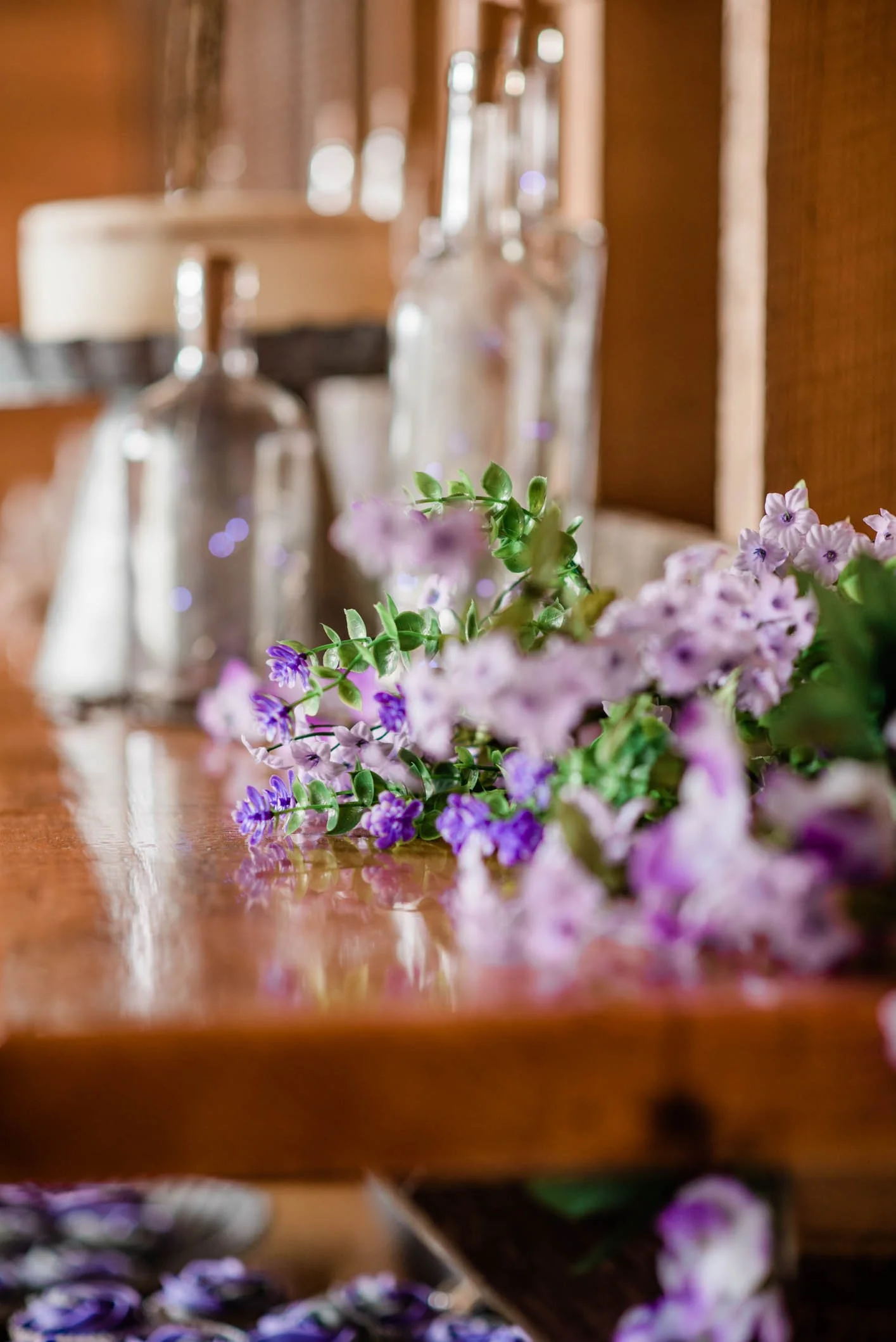 Close-up of purple and pink flowers on a wooden table with glass bottles and jars in the background.