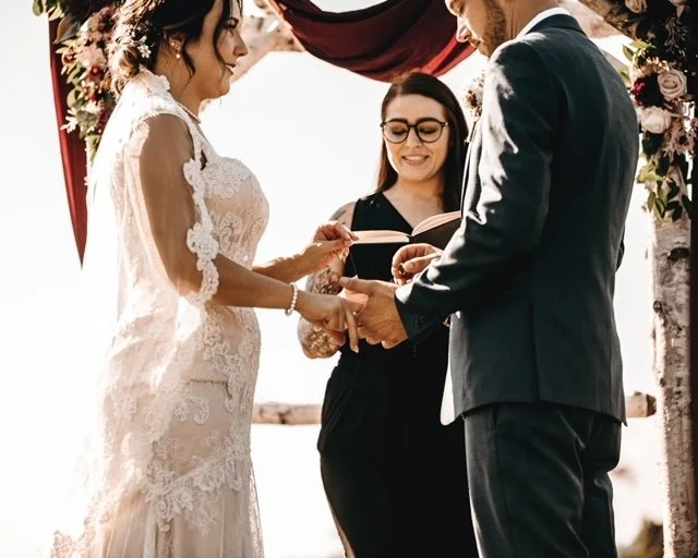 Bride and groom holding hands during wedding ceremony with officiant reading vows, outdoors with floral decorations.