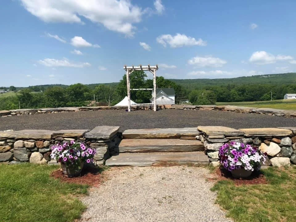 Stone steps leading to a flat area with a wooden frame in the distance, surrounded by flower pots with purple and white flowers, overlooking a green landscape with buildings and trees under a partly cloudy sky.