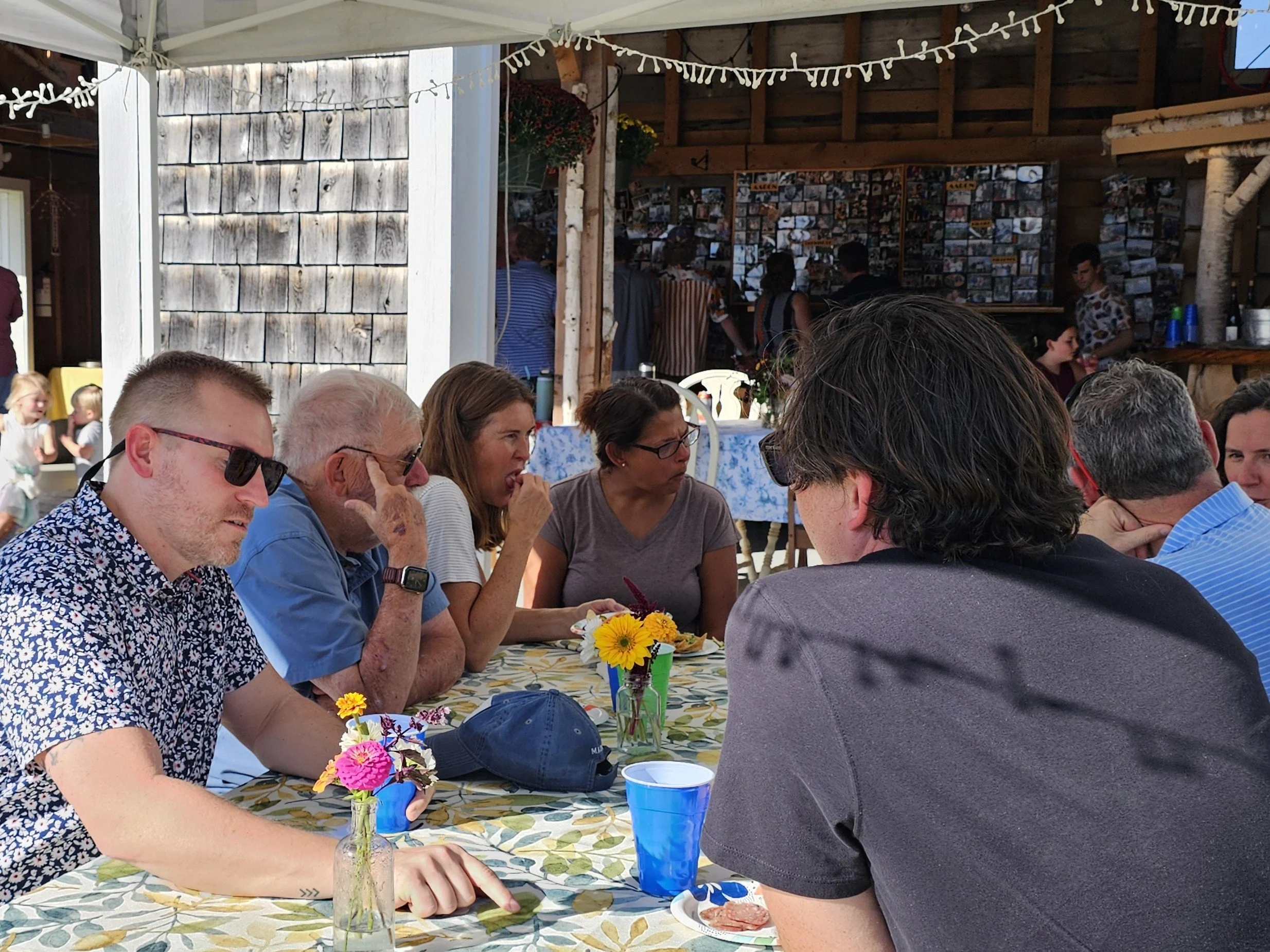 People sitting at a table with colorful flowers in small vases, engaged in conversation at an outdoor gathering under a canopy with string lights, in a rustic wooden setting.